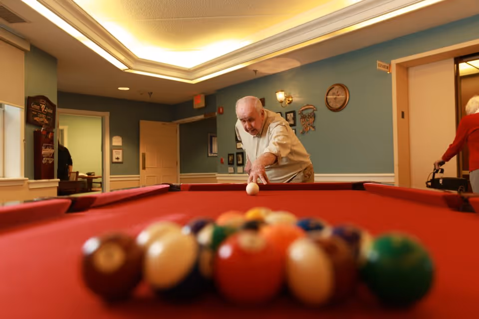 An elderly man is playing pool on a red felt pool table in a well-lit room with blue walls. In the background, an elderly woman with a walker is seen near an elevator. The room has framed pictures on the wall and a ceiling light fixture.