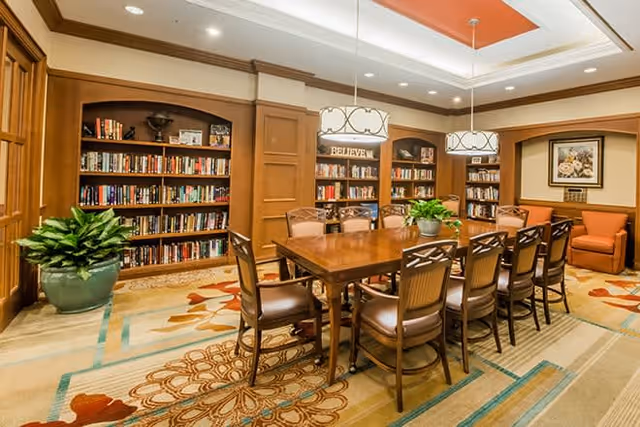 A well-lit library or reading room with wooden bookshelves filled with books along the walls. In the center, there is a large wooden table surrounded by ten chairs. Two hanging light fixtures illuminate the table. The room has a patterned carpet, a large potted plant, and a framed floral artwork on the wall. The ceiling features a recessed area with an orange accent.