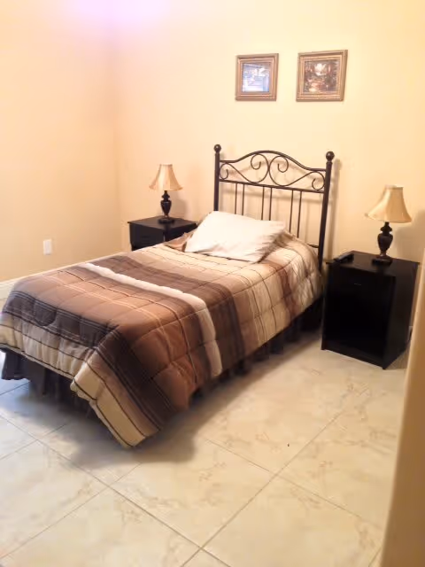 Small bedroom with a single bed featuring a metal headboard, plaid bedding, two nightstands with lamps, and two framed pictures on a beige wall.