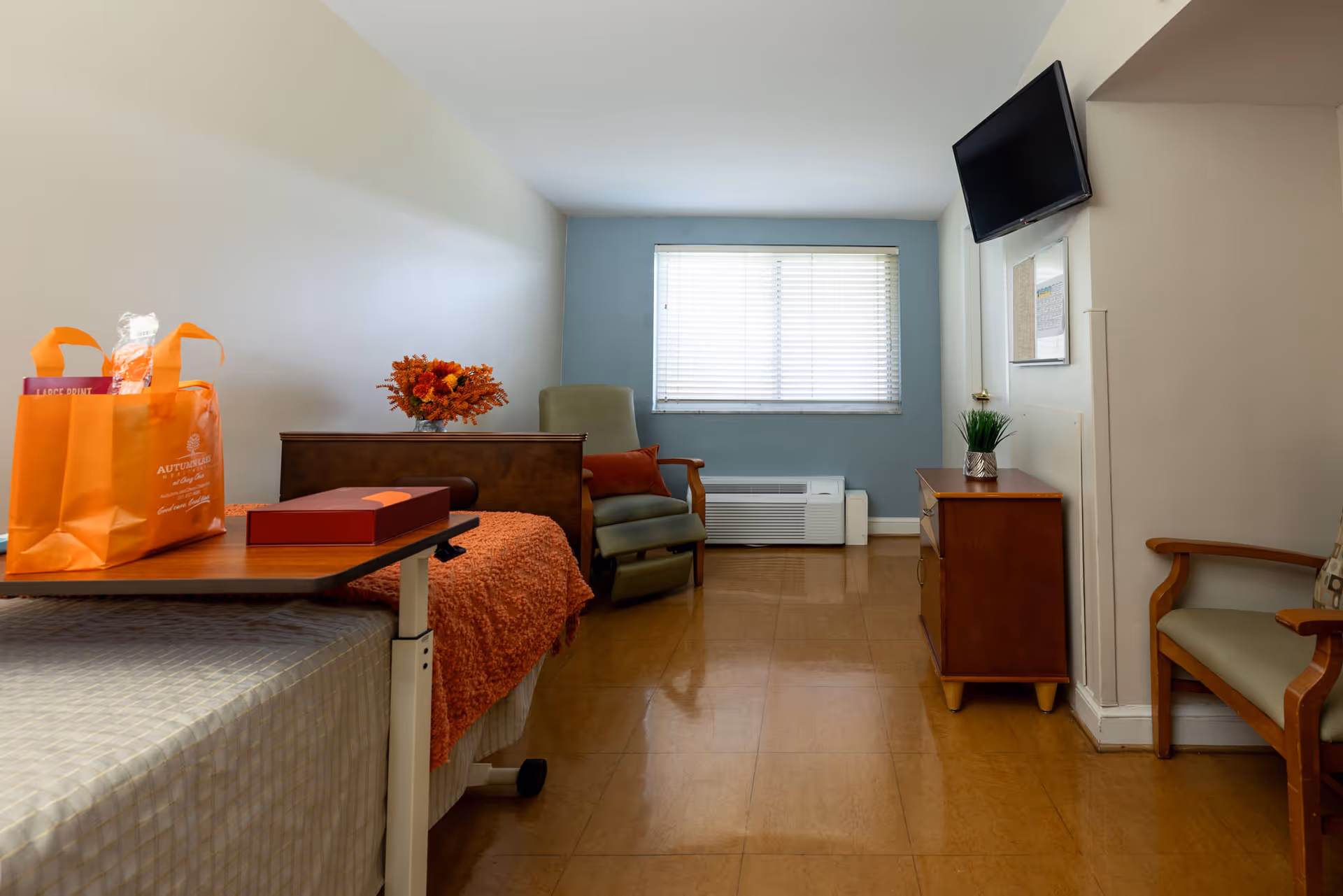 A simple, clean senior living bedroom with a bed covered in a light-colored blanket and an orange throw. On the bed's overbed table, there is an orange gift bag and a red box. The room has a green armchair with an orange pillow, a wooden dresser with a small plant, a wall-mounted TV, and a window with blinds letting in natural light. The floor is a polished brown tile.