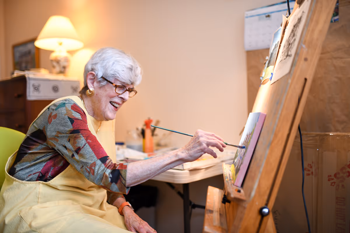 An elderly woman with white hair and glasses is smiling while painting on a canvas set on an easel in a cozy indoor room. She is wearing a colorful patterned shirt and a yellow apron. In the background, there is a table with art supplies and a lamp providing warm light.