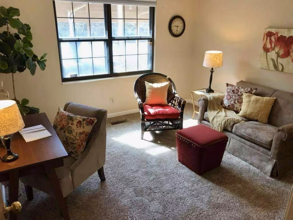 A cozy living room with a large window letting in natural light. The room features a gray sofa with decorative pillows and a throw blanket, a wicker chair with a red cushion and a white pillow, a red ottoman, a wooden desk with a floral lamp and a notebook, a gray upholstered chair with a floral pillow, a side table with a lamp, a wall clock, a painting of red flowers, and a green potted plant in the corner.