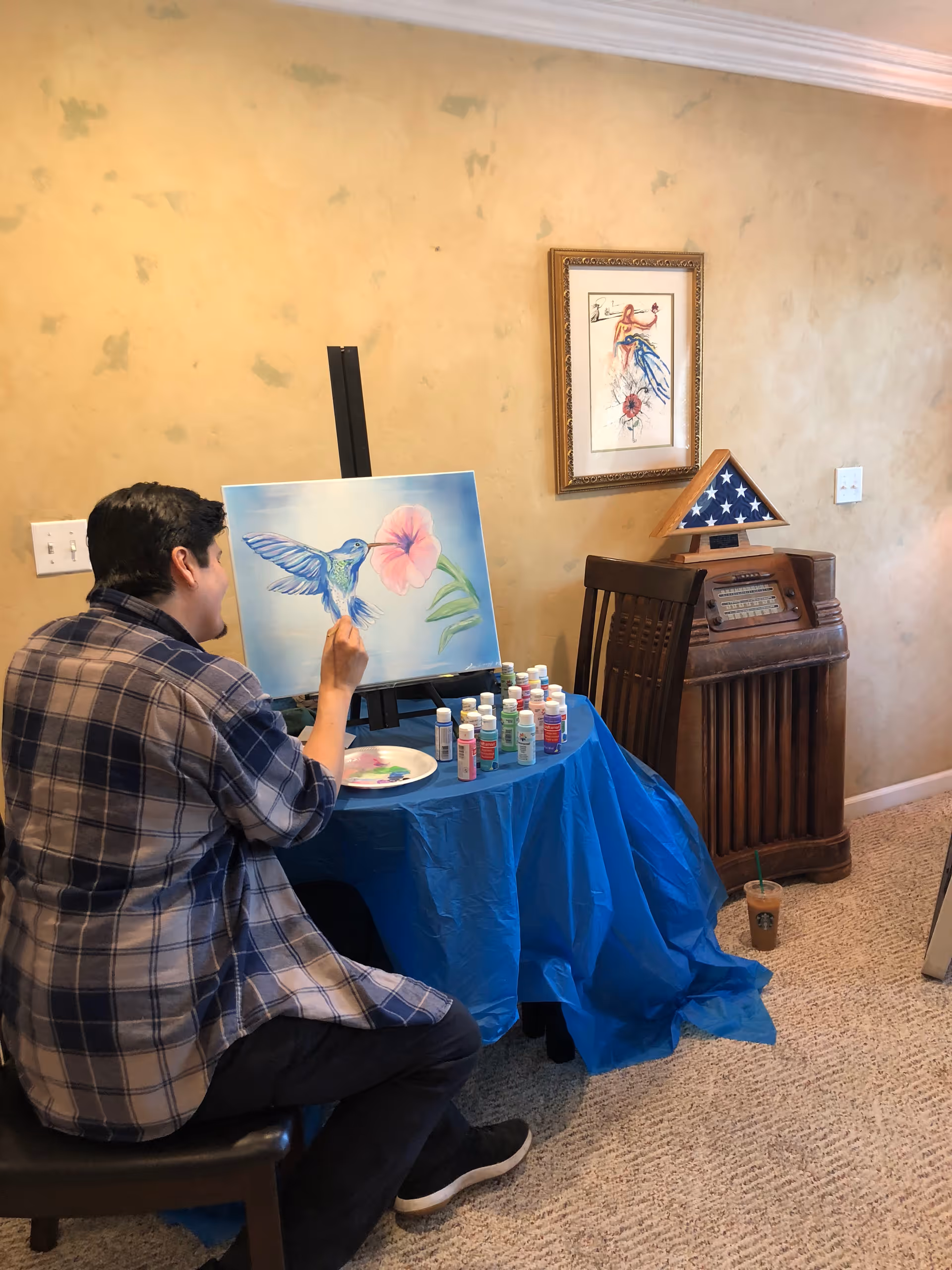 A person seated on a chair painting a colorful hummingbird and flower on a canvas placed on an easel. The table in front of them is covered with a blue cloth and holds several bottles of paint and a paint palette. The room has beige textured walls, a framed artwork, a wooden cabinet with a folded American flag in a triangular display case, and a carpeted floor.