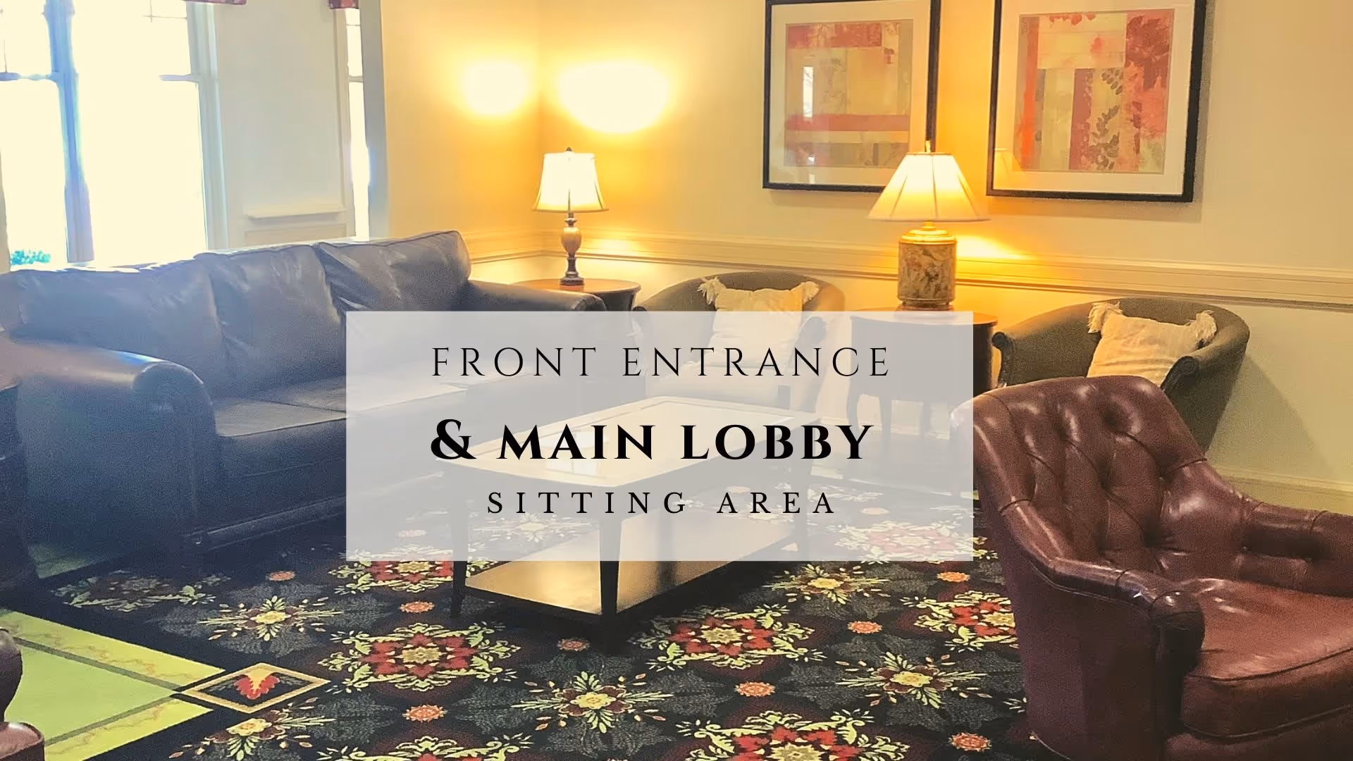 A cozy sitting area in the front entrance and main lobby of a facility, featuring a dark leather sofa, two armchairs with cushions, a coffee table, two table lamps, and framed artwork on the wall. The floor is covered with a floral patterned carpet.
