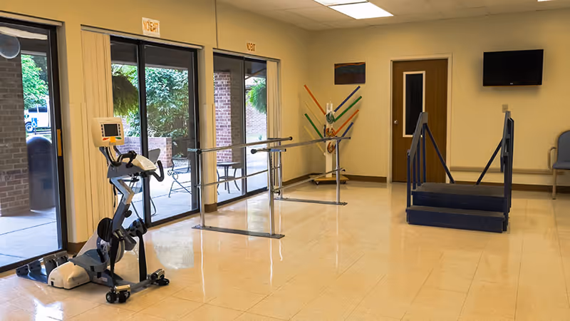 A rehabilitation room with exercise equipment including a stationary bike, parallel bars for walking practice, and a small set of stairs with handrails. The room has large windows and glass doors leading outside, beige walls, and a mounted TV on the wall.