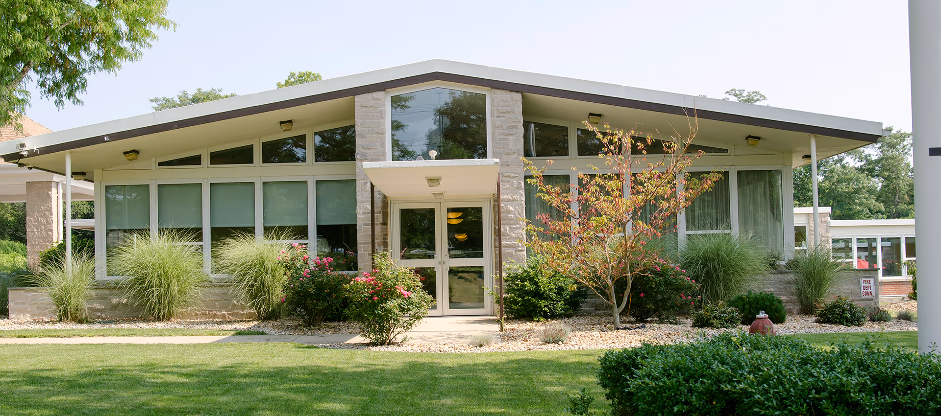 Exterior view of a single-story building with large windows and a slanted roof, surrounded by green grass, bushes, and small trees under a clear sky.