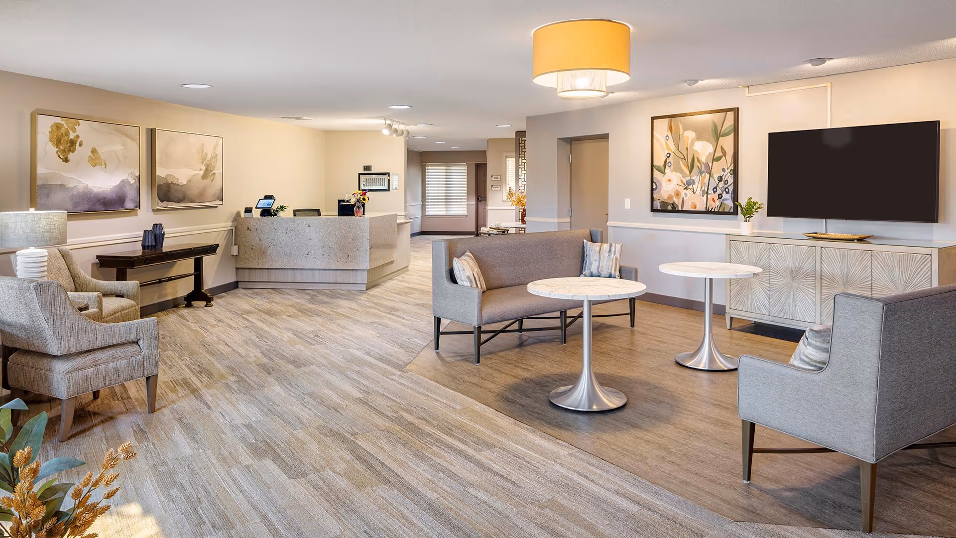 A spacious and well-lit senior living facility common area with modern furniture including armchairs, a loveseat, and round tables. The room features light-colored walls adorned with abstract and floral artwork, a large flat-screen TV mounted above a decorative cabinet, and a reception desk in the background. The flooring is a mix of wood and carpet, and a ceiling light fixture provides warm illumination.