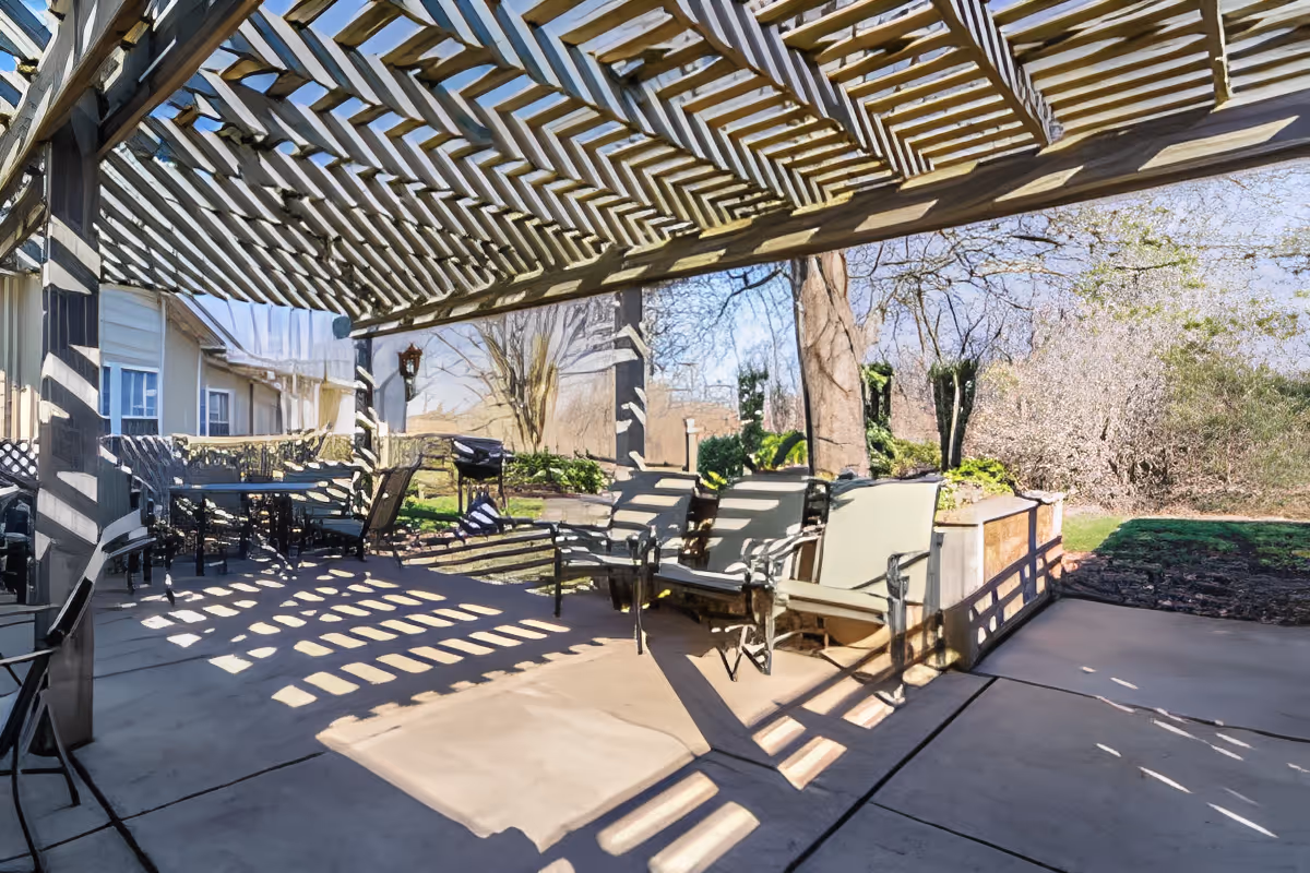 Shaded outdoor patio with chairs and tables under a wooden pergola overlooking a landscaped yard.