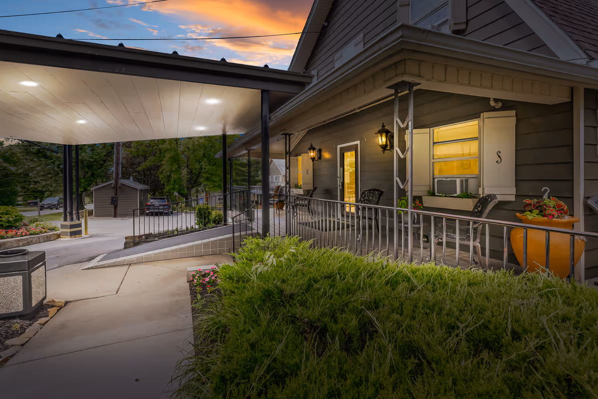 Covered front entrance and porch of a senior living facility with a wheelchair ramp, outdoor seating, and potted plants at dusk.