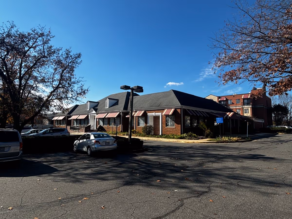 Low-rise brick senior residence building with striped awnings, parked cars, and trees under a clear blue sky.