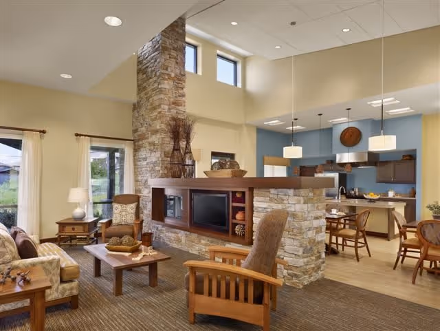 A spacious and well-lit living room area with a stone fireplace and built-in shelving. The room features comfortable wooden furniture including armchairs and a sofa, a coffee table, and side tables with lamps. Large windows with curtains allow natural light to enter. In the background, there is a dining area with tables and chairs, and a kitchen with modern cabinetry and pendant lighting.