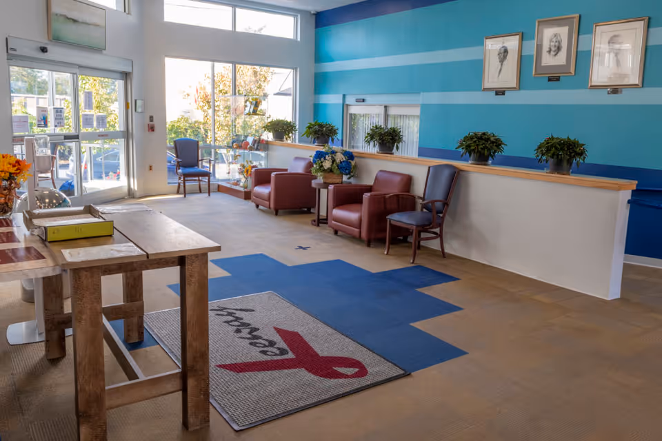 A bright and welcoming facility lobby with large windows letting in natural light. The room features a wooden table with papers and a binder, a mat with a red ribbon and the word 'Leeway', several chairs including two red armchairs and a blue chair, potted plants on a half wall, and framed portraits on a blue striped wall.