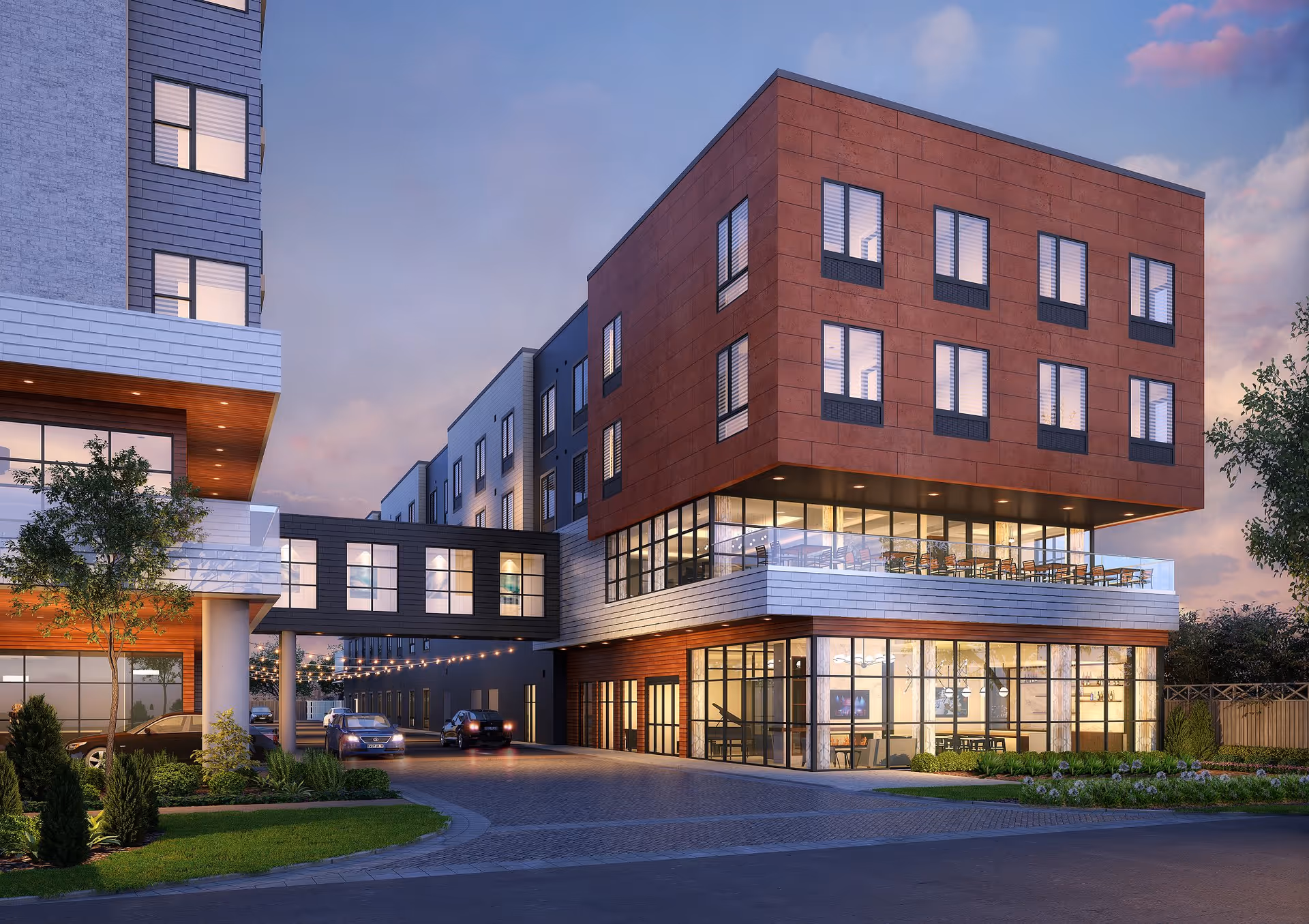 Exterior view of a modern multi-story building at dusk with large windows, a skybridge connecting two sections, cars parked along the driveway, and landscaped greenery around the entrance.