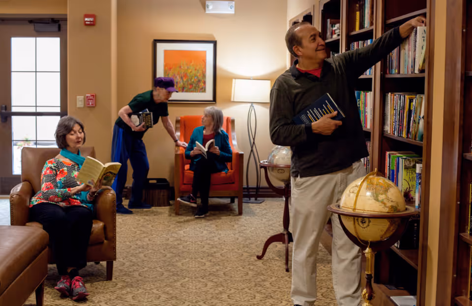 Four elderly people in a cozy library or reading room. One woman sits in a leather chair reading a book, a man stands near a bookshelf selecting a book, another woman sits in an orange armchair reading, and an elderly man with a purple hat is bending down holding a book. The room has warm lighting, a patterned carpet, and two globes on stands.