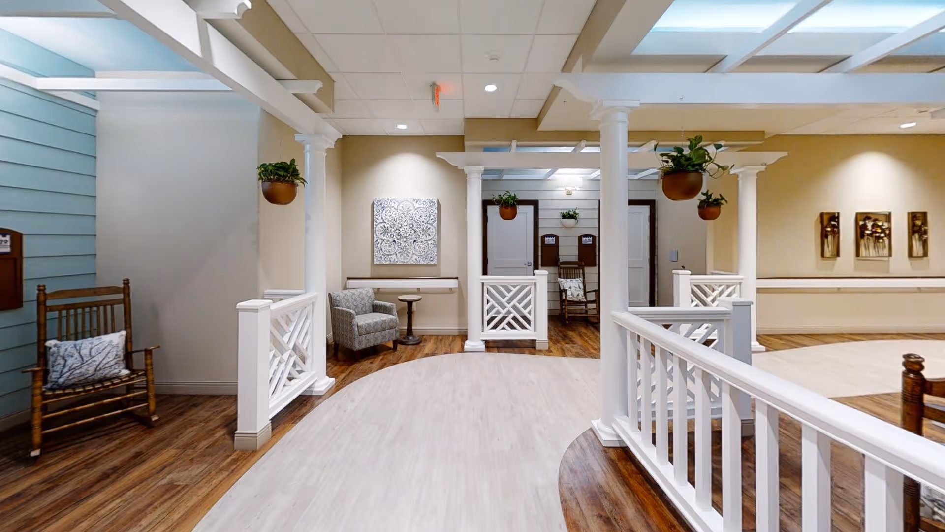 Interior view of a memory care facility hallway with wooden rocking chairs, cushioned armchairs, hanging plants, white columns, and decorative wall art. The floor has a combination of light and dark wood finishes, and the space is well-lit with ceiling lights.