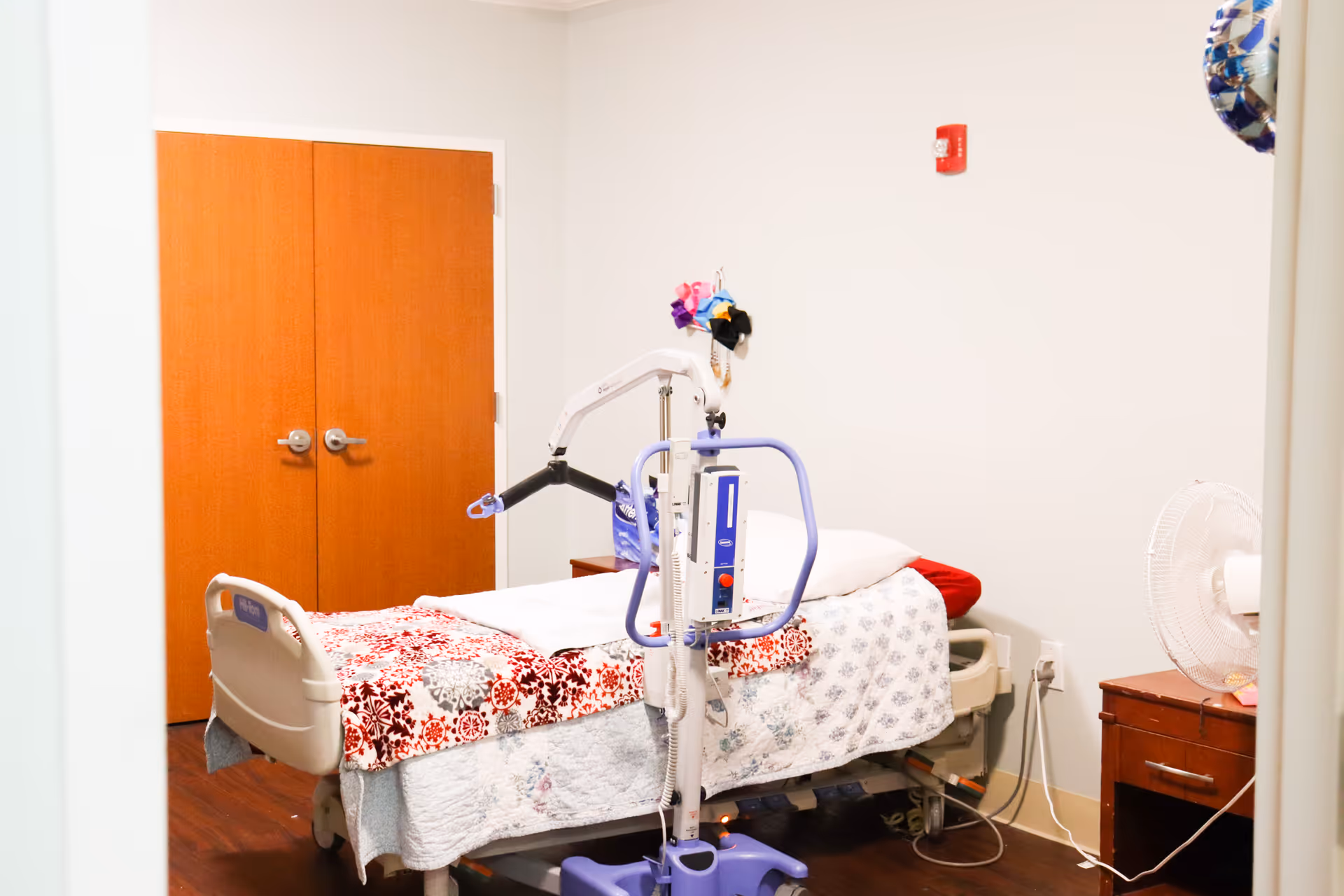 A small room with a hospital bed covered with a patterned quilt and a red pillow. There is a patient lift device positioned next to the bed. The room has wooden double doors, a wooden nightstand with a white fan on it, and a white wall with a fire alarm mounted on it.