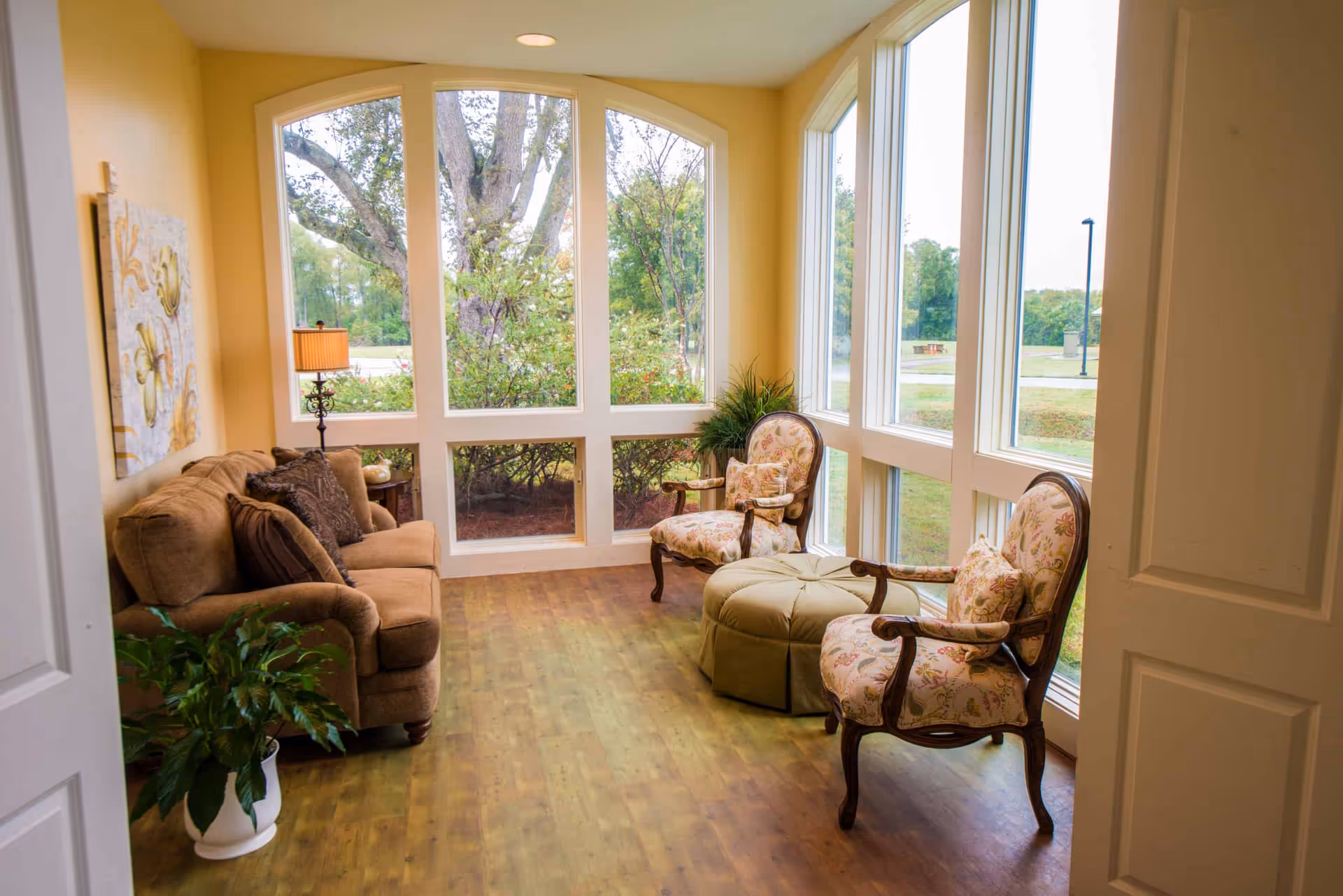 Bright sunroom-style sitting area with a sofa and two upholstered chairs around an ottoman facing large floor-to-ceiling windows.