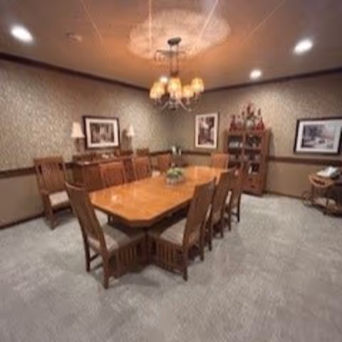 A formal dining room with a long wooden table surrounded by eight wooden chairs with cushioned seats. The room has beige walls with a decorative chair rail, two framed pictures, a wooden cabinet with decorative items, and a chandelier hanging from the ceiling. The floor is carpeted in a light gray color.