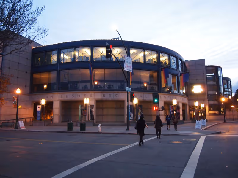 Exterior view of a modern, curved building at dusk with large glass windows and illuminated interior lights. Several people are crossing the street in front of the building, and street lamps and traffic lights are visible.