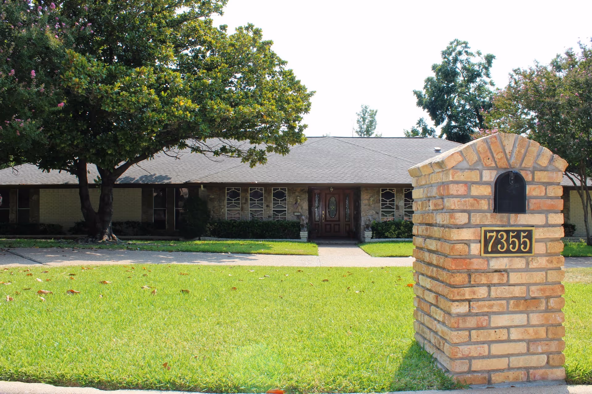 Front exterior view of a single-story brick building with a gray shingled roof, surrounded by green grass and trees. In the foreground, there is a brick mailbox with the number 7355 displayed on it.
