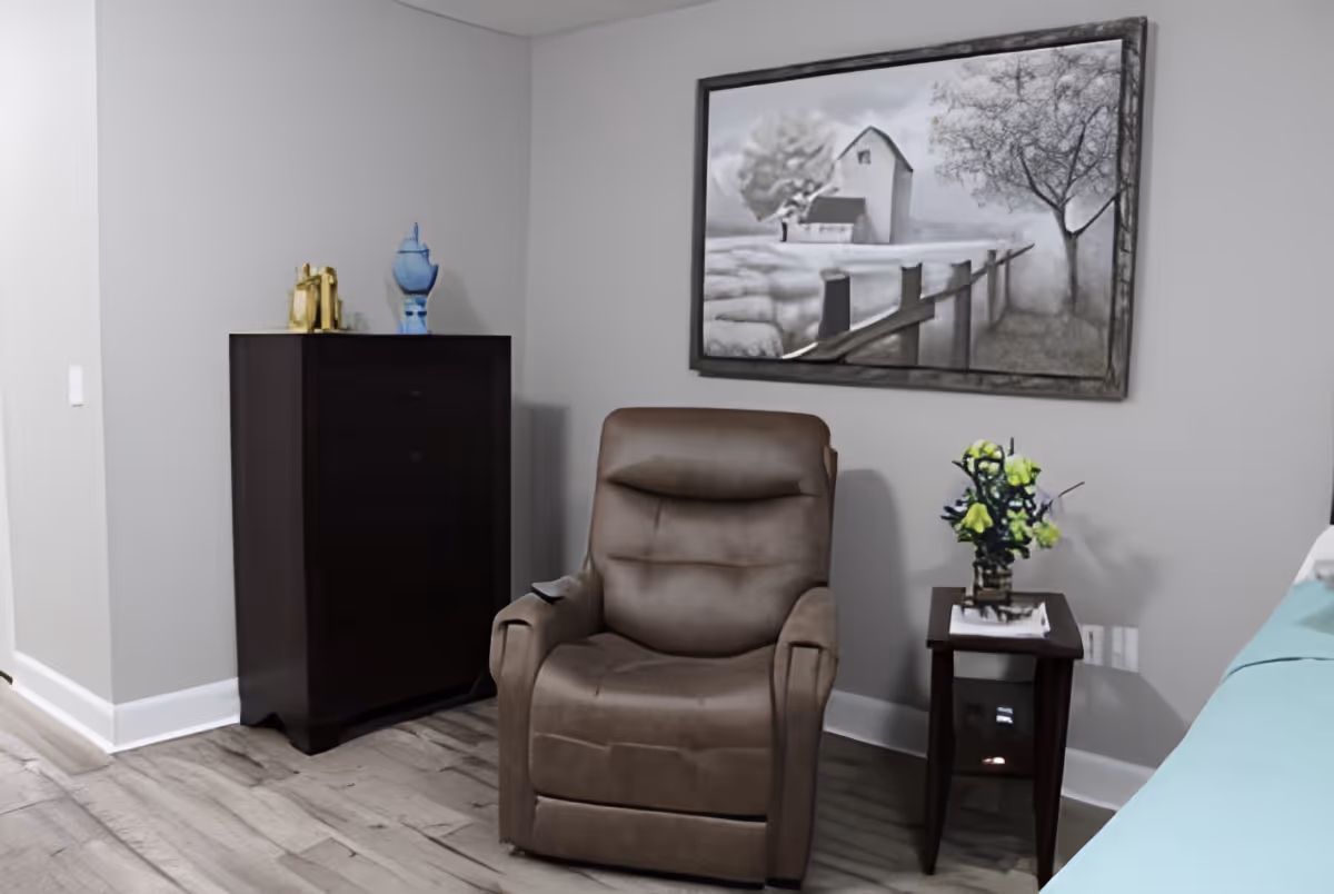 A cozy corner of a room featuring a brown recliner chair, a small dark wooden side table with a vase of yellow flowers, a dark wooden chest of drawers with decorative items on top, and a large framed black and white painting of a barn and fence on the wall.