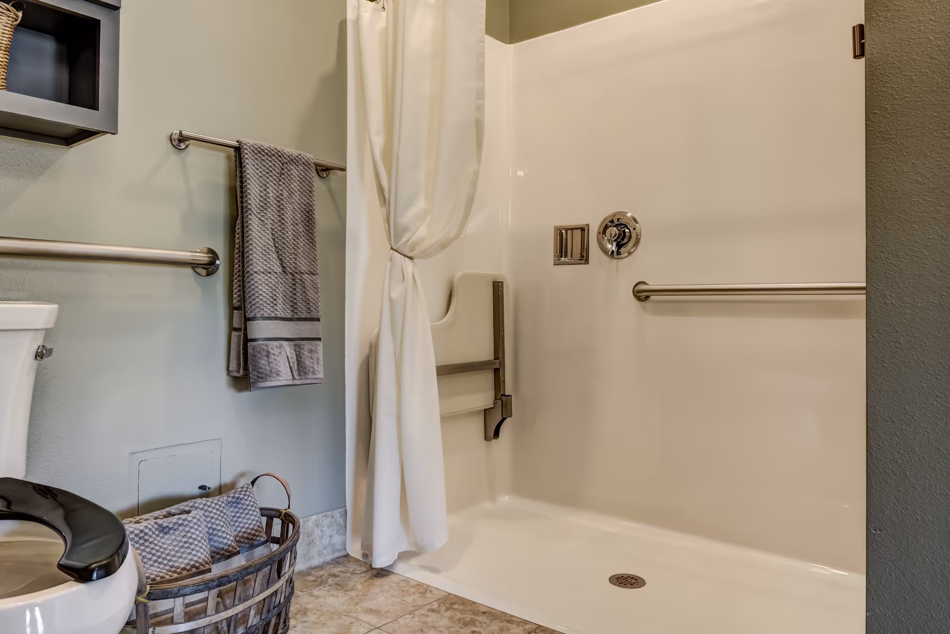 A bathroom featuring a walk-in shower with a white curtain tied to the side, a foldable shower seat, and stainless steel grab bars. There is a towel rack with a gray towel hanging on it, a toilet with a black seat, and a basket on the floor containing folded towels. The walls are painted light green and the floor has beige tiles.