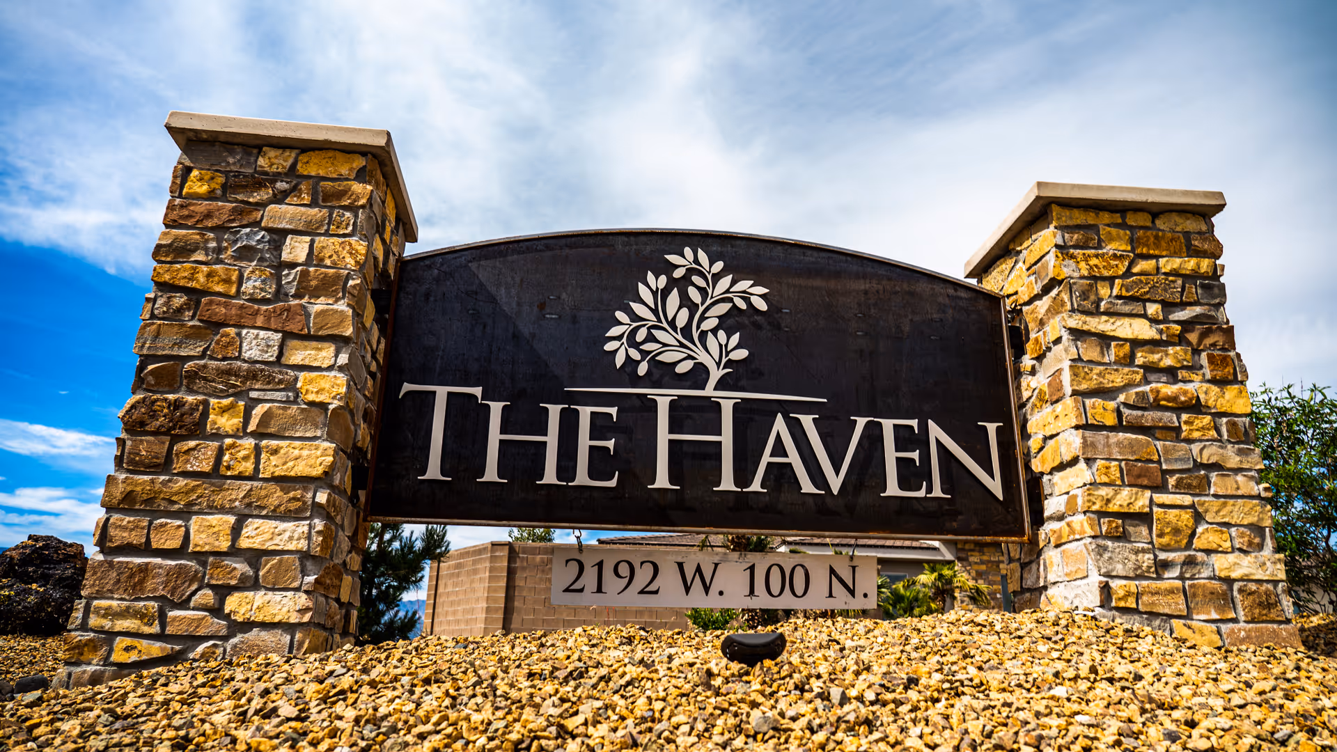 Stone monument sign with two stone pillars on each side and a black plaque in the center displaying the text 'THE HAVEN' with a tree logo above it and the address '2192 W. 100 N.' below. The sky is partly cloudy in the background.