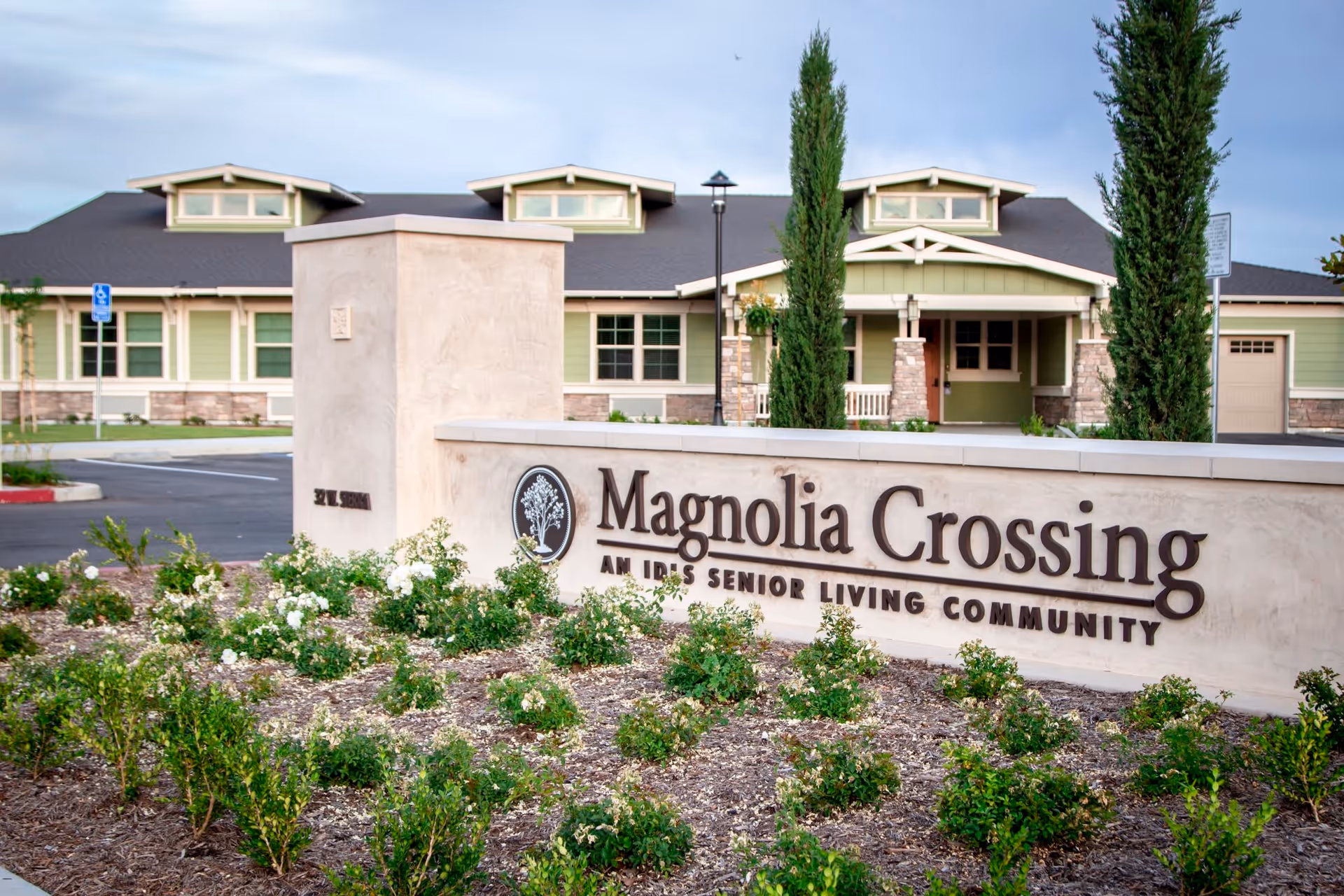 Entrance sign for Magnolia Crossing, an IDES senior living community, with a landscaped garden in front and a large building with green siding and stone accents in the background.