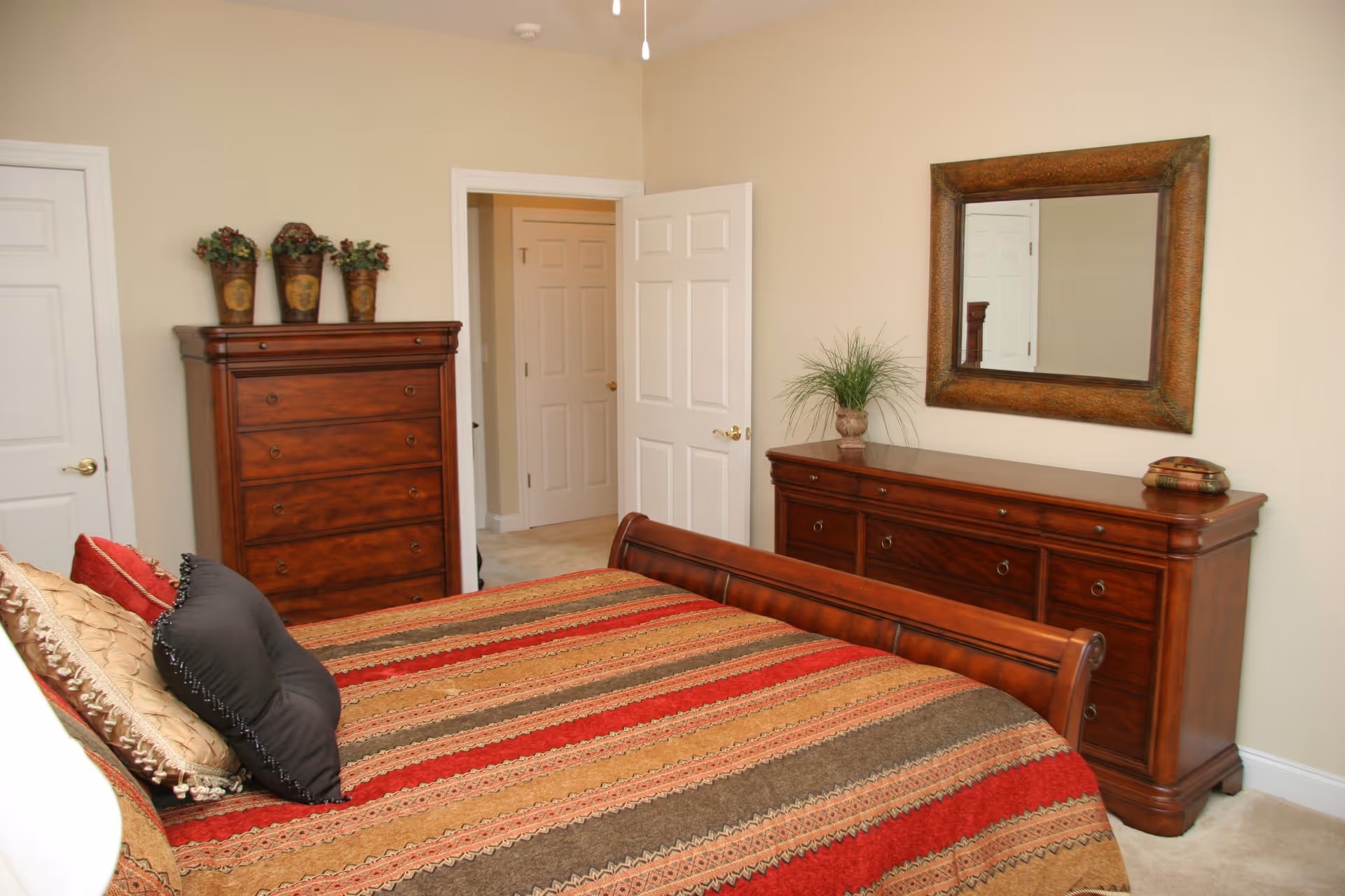 A bedroom with a wooden bed frame and a multicolored striped bedspread in shades of red, brown, and beige. There are several pillows on the bed. The room also features a wooden dresser with a large framed mirror above it, a tall wooden chest of drawers with three decorative plant pots on top, beige walls, and two white doors, one of which is open showing a hallway.