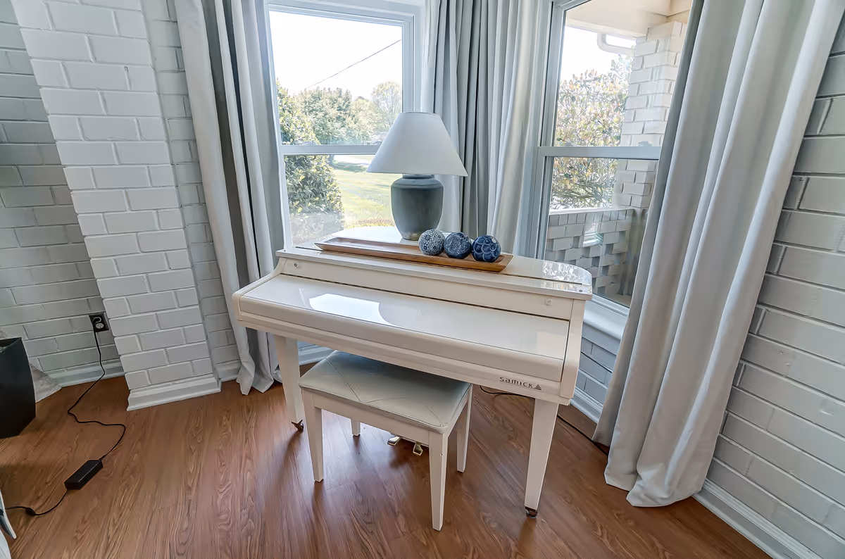 A white upright piano with a matching white bench placed in front of large windows with light gray curtains. On top of the piano is a blue-gray lamp and a wooden tray holding three decorative blue and white spheres. The room has white painted brick walls and wood flooring.