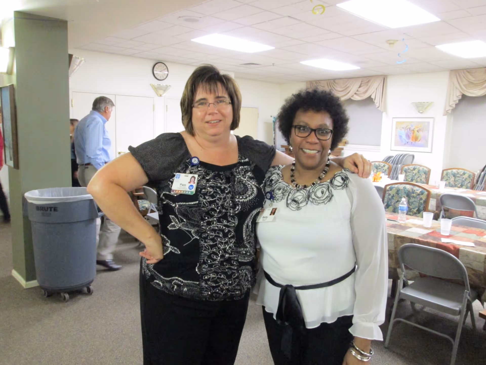 Two women standing indoors in a room with tables and chairs, smiling at the camera. Both women are wearing name badges and glasses. The room has a clock on the wall, a trash bin, and some people in the background.