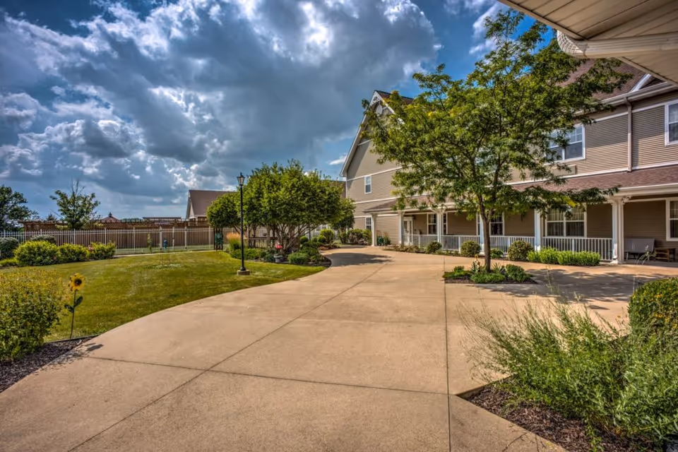 Courtyard and paved walkway leading to a two-story senior living building with trees, lawn, and a dramatic cloudy sky.