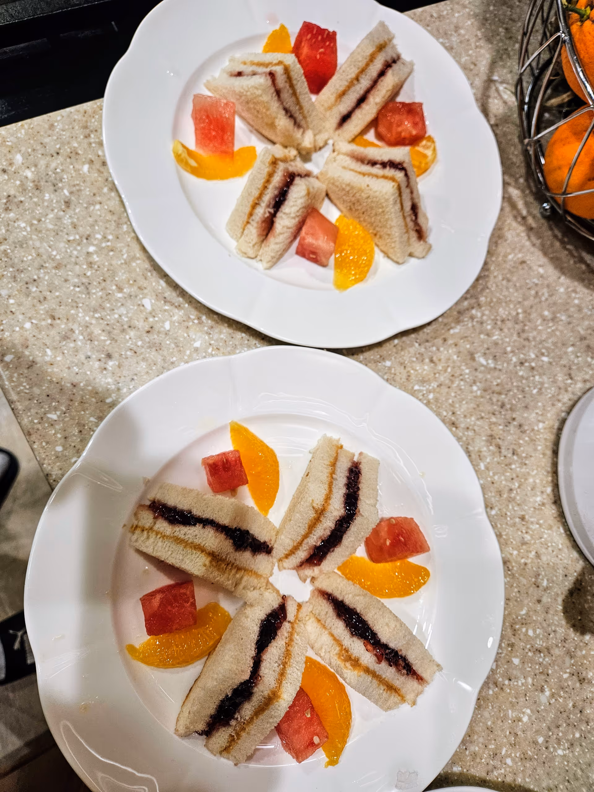 Two white plates holding triangular jam sandwiches garnished with orange slices and small watermelon cubes on a speckled countertop.