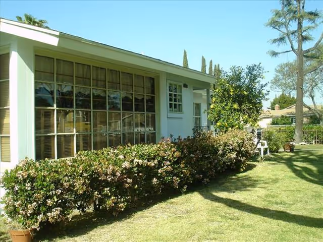 Exterior view of a single-story building with large windows and a well-maintained garden featuring flowering bushes and a tree. The lawn is green and there is a white chair near the building.