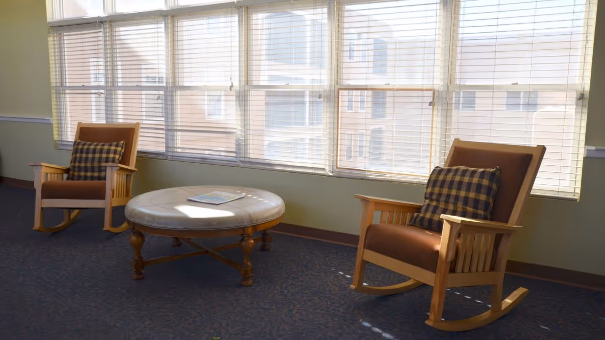 A bright sitting area with two wooden rocking chairs featuring brown cushions and plaid pillows, positioned around a round wooden coffee table with a light-colored top. Large windows with blinds allow natural light to fill the room.