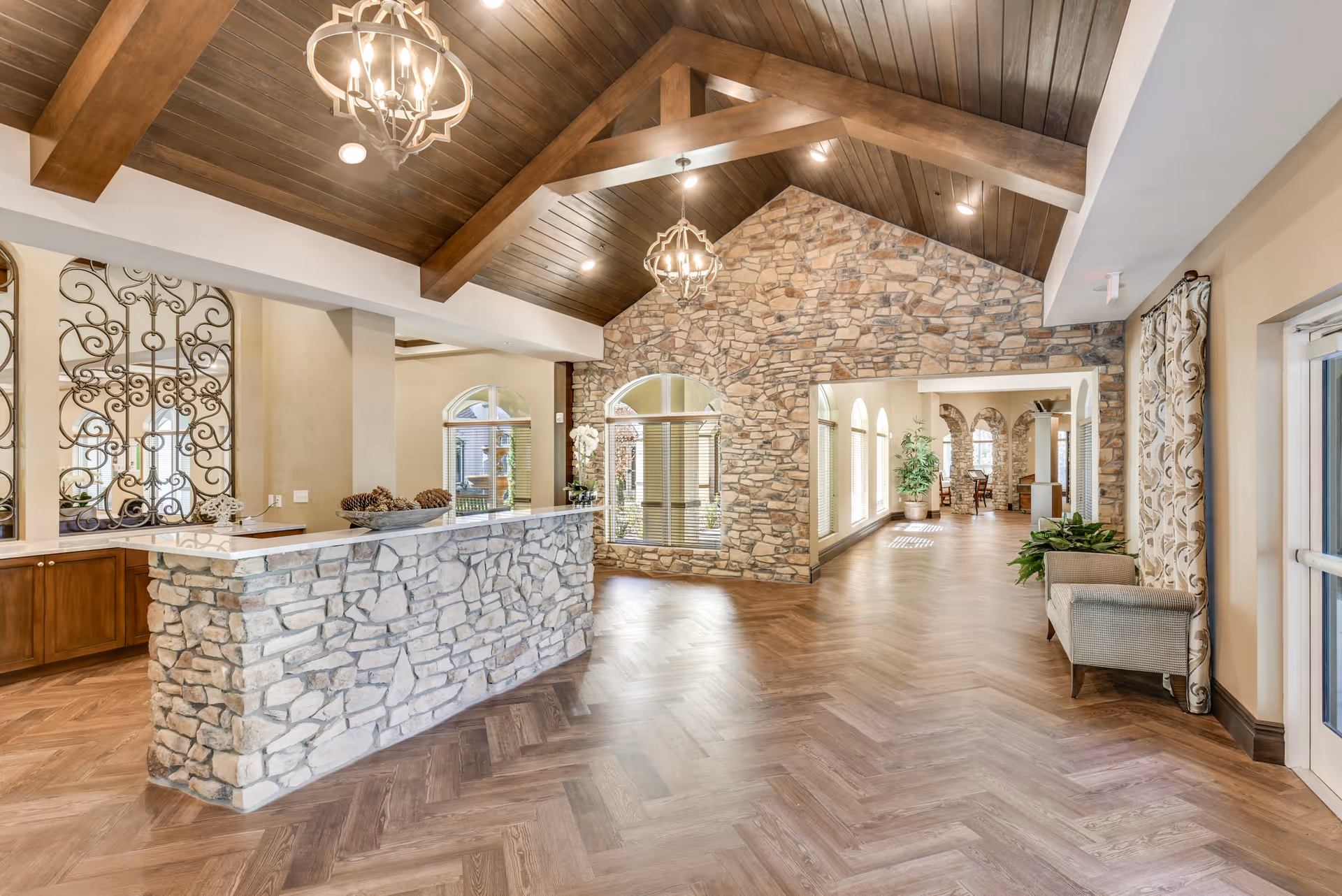 Interior view of a senior living facility featuring a stone reception desk with decorative wrought iron panels behind it, wooden vaulted ceiling with exposed beams and chandeliers, stone accent wall with arched windows, wood-patterned flooring, and a seating area with a cushioned bench and plants near the entrance.