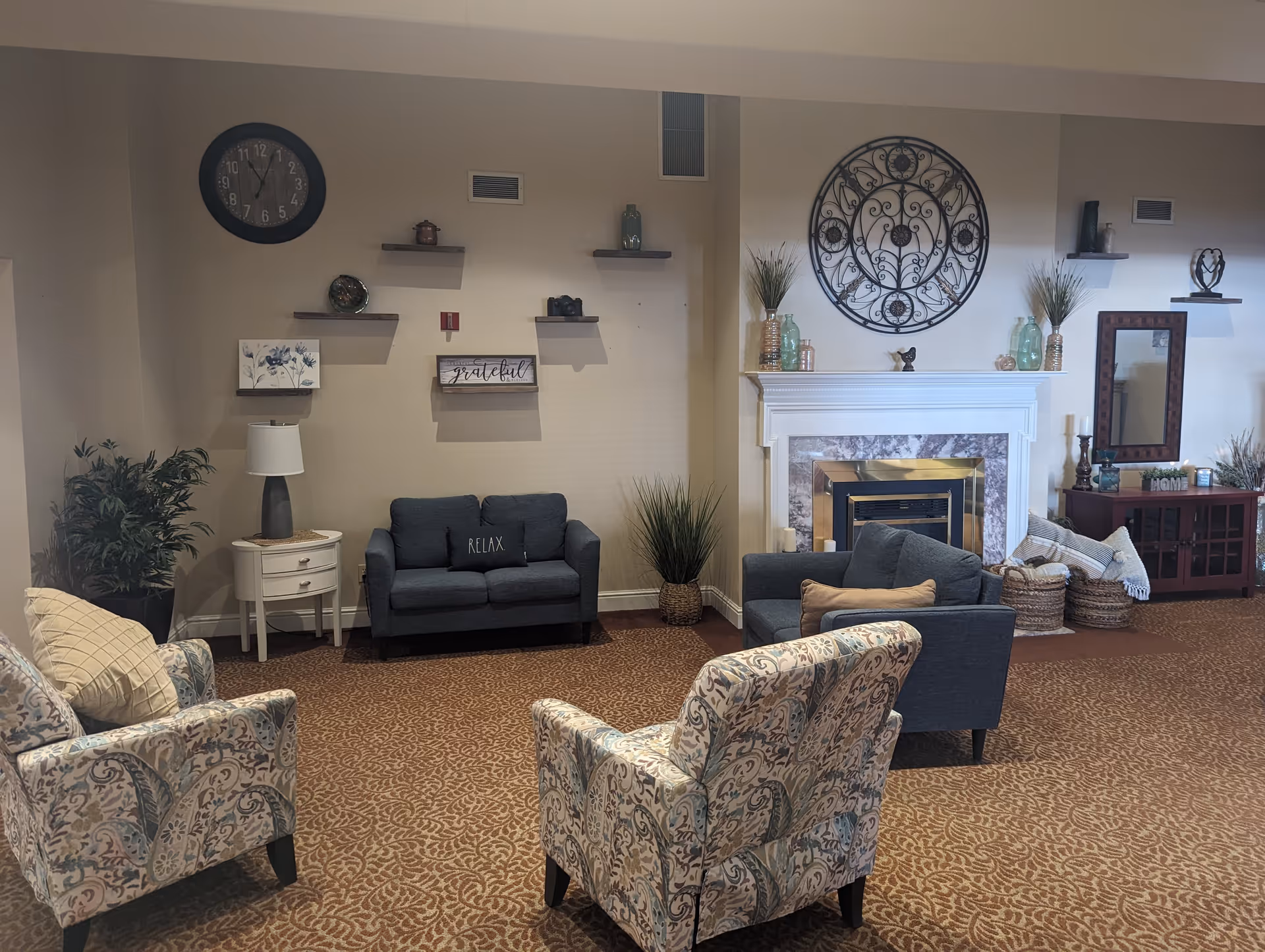 A cozy living room area with patterned carpet, two floral armchairs, a small dark blue loveseat with a 'RELAX' pillow, and a matching armchair. The room features a white fireplace with decorative vases and plants on the mantel, a large ornate circular wall decoration above the fireplace, and several floating shelves with small decor items. There is a side table with a lamp and a large wall clock, as well as a wooden cabinet with a mirror and more decorative items. The walls are painted beige.