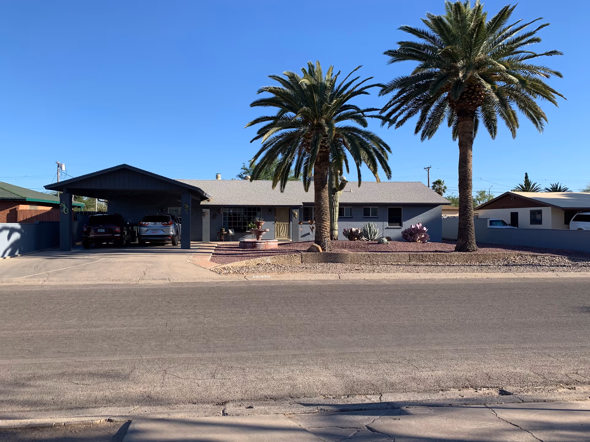 Exterior front view of a single-story residential building with a covered carport housing two vehicles. The yard features desert landscaping with gravel, two tall palm trees, and various desert plants. The sky is clear and blue.