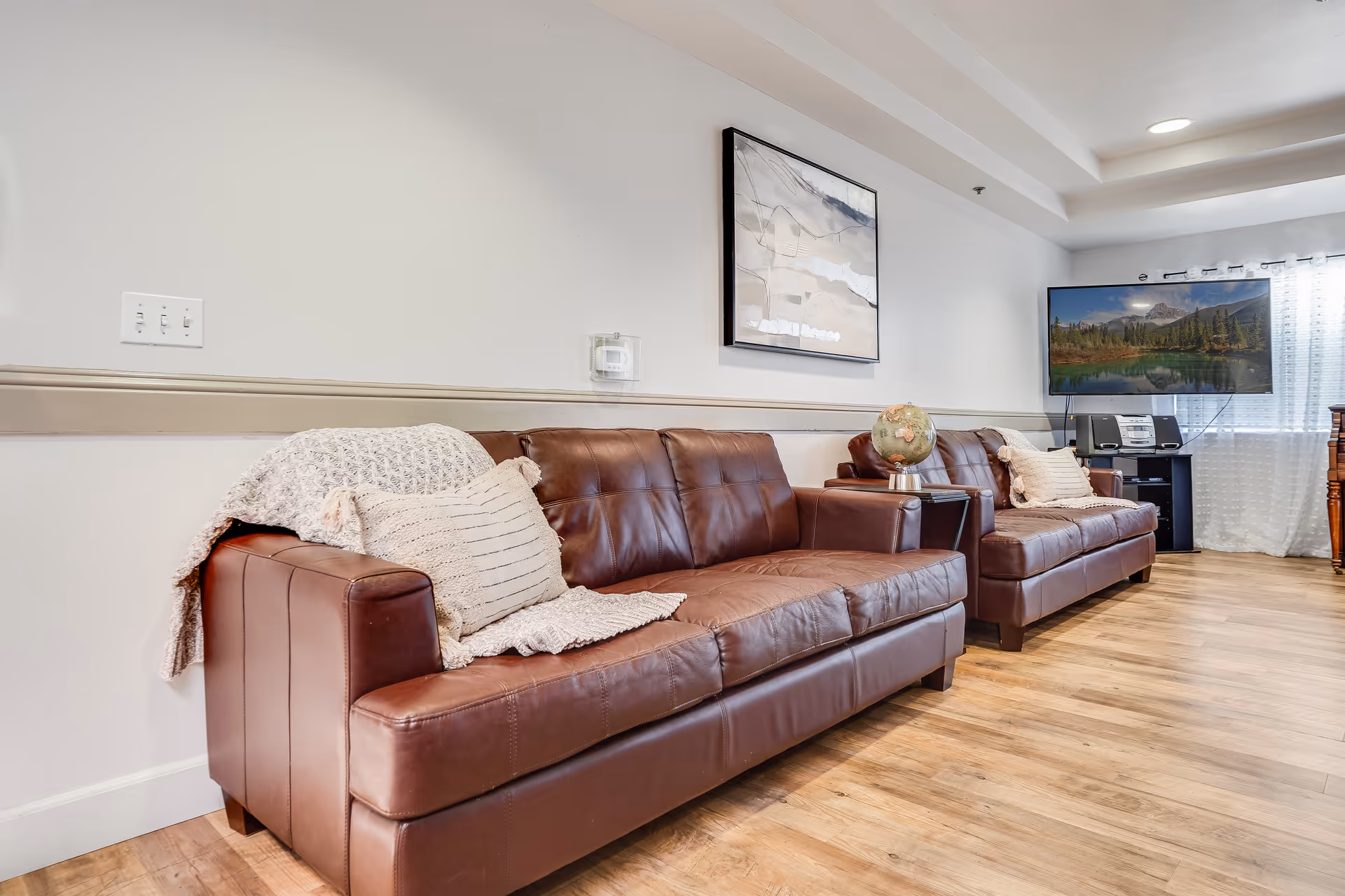 Interior of a senior living facility lounge area with two brown leather sofas adorned with beige knitted throws and pillows. A small side table between the sofas holds a globe. A large flat-screen TV is mounted on the wall near a window with sheer white curtains, and a piece of abstract art hangs above the sofas. The room has wooden flooring and white walls with a beige chair rail.