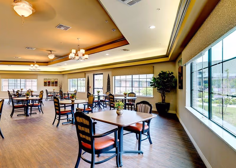 A bright and spacious dining room with multiple wooden tables and chairs arranged neatly. Each table has a small decorative plant. Large windows on the right side allow natural light to fill the room, and the ceiling features recessed lighting and ceiling fans. The floor is wooden, and the walls are painted in a light neutral color.