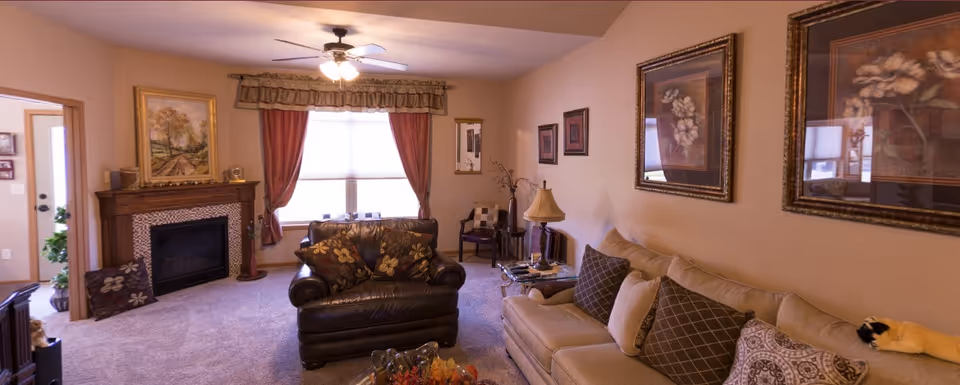 A cozy living room in a retirement community featuring a beige sofa with multiple patterned cushions, a dark brown leather armchair, a wooden fireplace with a framed painting above it, a window with red curtains and a valance, a ceiling fan with lights, and framed floral artwork on the walls.