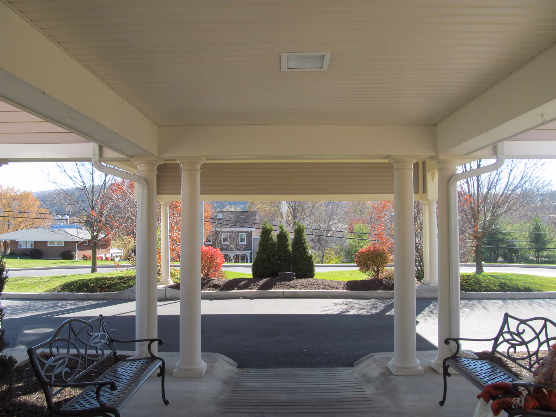 View from a covered entrance area supported by white columns, looking out onto a driveway and landscaped area with bushes and trees showing autumn colors. Two black metal benches with decorative backs are placed on either side of the entrance.