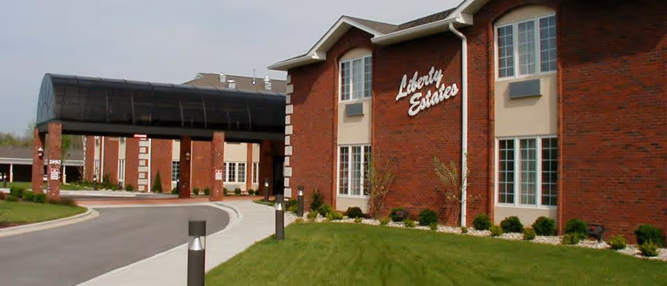 Exterior view of a senior living facility named Liberty Estates, featuring a red brick building with large windows and a covered driveway entrance. The surrounding area includes a curved road, green grass, and small shrubs along the building.