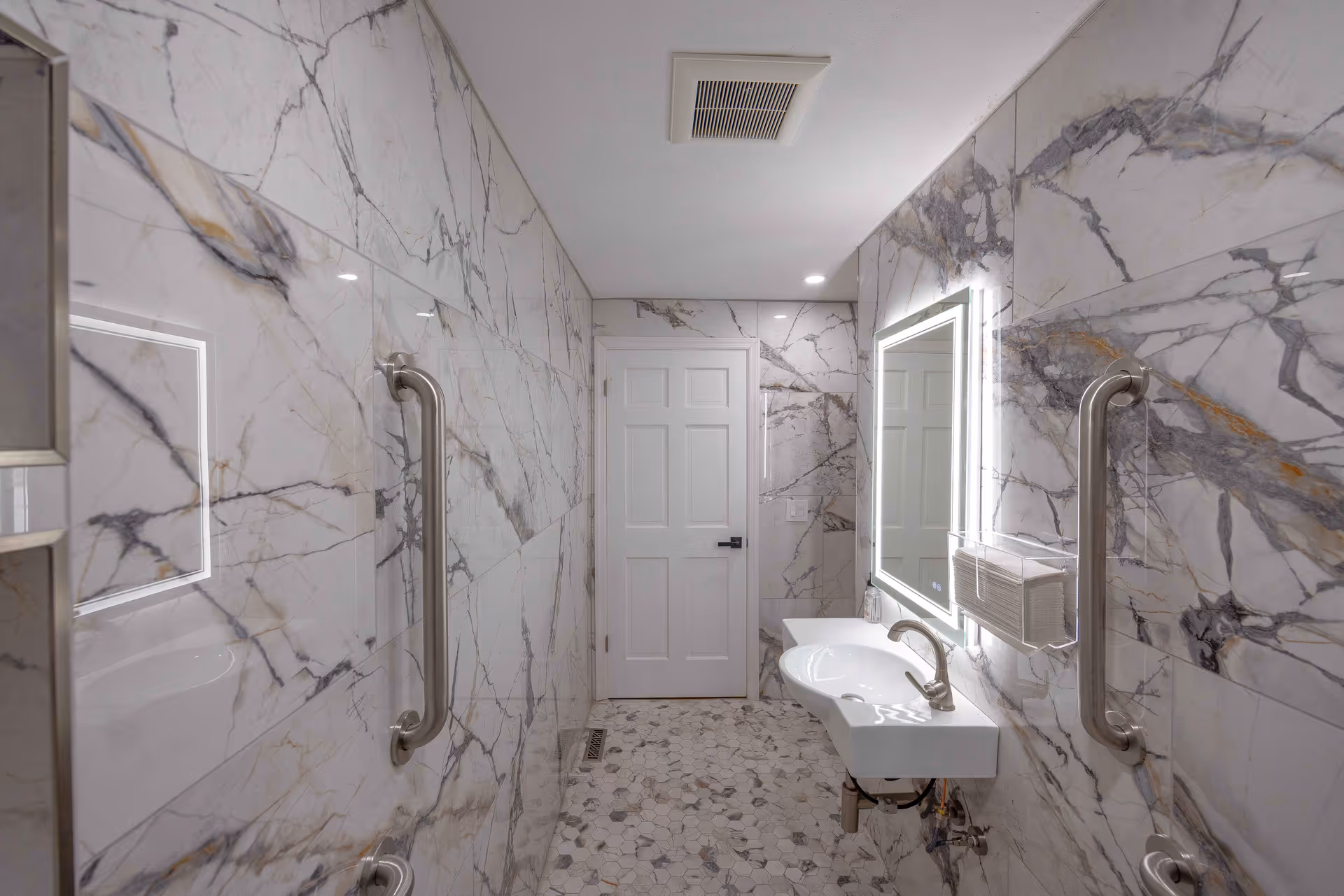 A modern bathroom with marble-patterned walls and floor tiles. The room features a white door at the end, a wall-mounted sink with a sleek faucet, a large illuminated mirror above the sink, and stainless steel grab bars on both sides of the walls for support.
