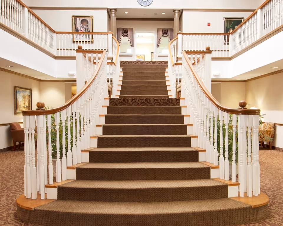 Wide carpeted staircase with white wooden railings and wooden handrails in a well-lit interior space, with paintings on the walls and seating areas on both sides.