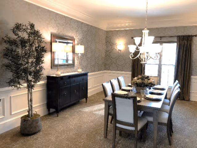 A well-lit dining room with a rectangular dining table set for eight people. The table has a floral centerpiece and place settings with plates and glasses. The room features patterned wallpaper, white wainscoting, a large mirror above a black sideboard, a potted plant in the corner, and a chandelier hanging above the table. There are large windows with curtains allowing natural light into the room.