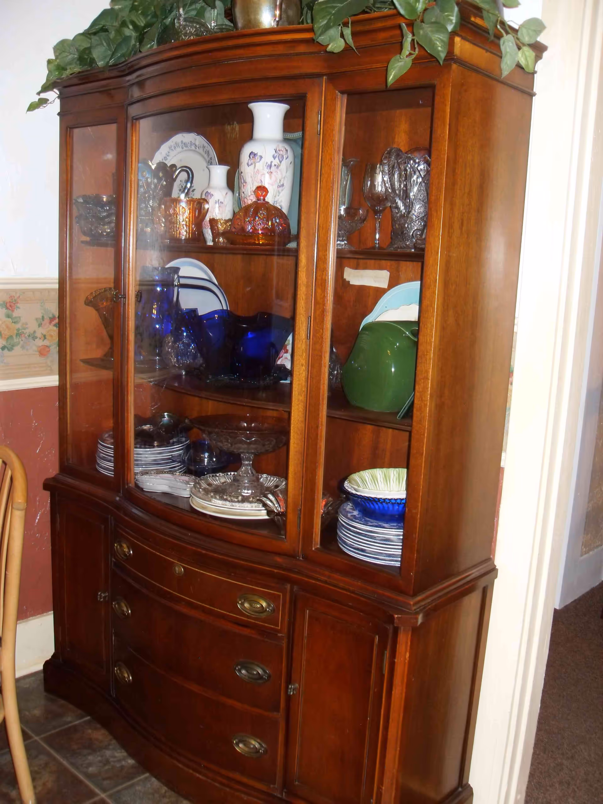 A wooden china cabinet filled with various glassware, plates, bowls, and decorative vases. The cabinet has glass doors on the upper section and drawers and cupboards on the lower section. There is a leafy green plant on top of the cabinet.