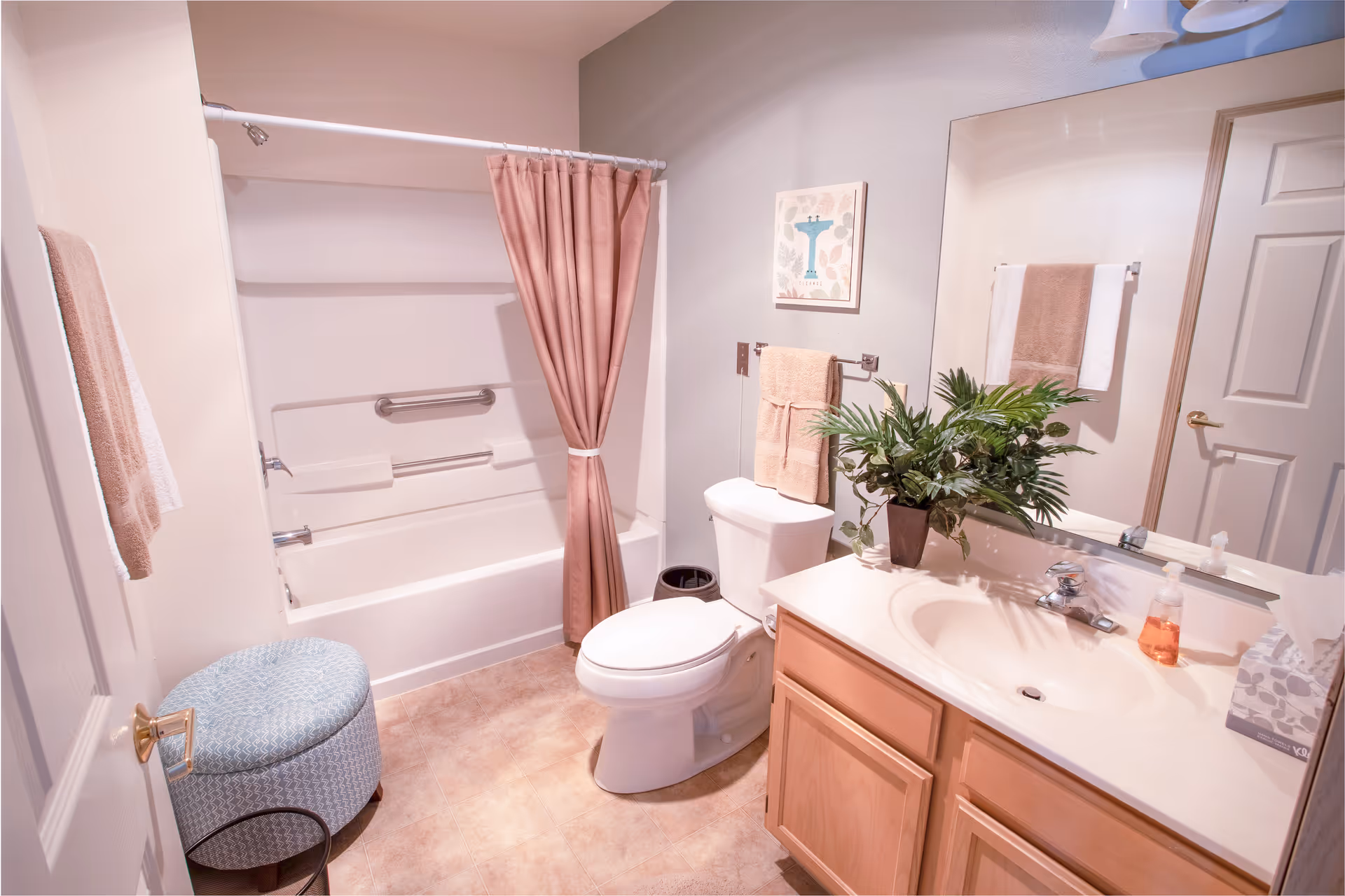 A clean and well-lit bathroom featuring a white bathtub with a pink shower curtain, a white toilet with beige towels hanging above it, a wooden vanity with a sink, a large mirror, a green potted plant on the counter, a blue patterned ottoman, and beige tiled flooring.