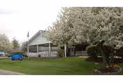 Exterior view of a single-story building with a covered porch, surrounded by green grass and blooming trees. An American flag is visible near the entrance, and a blue sign is placed on the lawn.