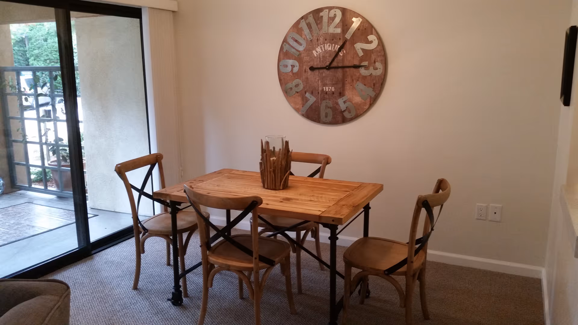 A small dining area with a wooden table and four matching wooden chairs with black metal supports. A large round wall clock with rustic design hangs on the beige wall above the table. To the left, there is a sliding glass door leading to an outdoor patio area with plants visible outside.